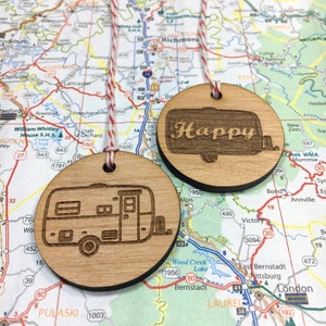 May include: Two wooden ornaments shaped like a camper van. One ornament has the word "Happy" engraved on it. The ornaments are hanging from a string against a map background.