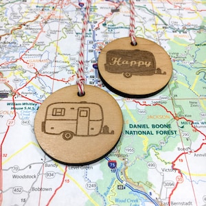 May include: Two wooden ornaments with a string attached. One ornament has a camper van design and the other has the word "Happy" with a camper van design. Both ornaments are on a map background.