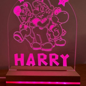 May include: A pink acrylic night light with a wood base. The light features a silhouette of Mario, Luigi, and Yoshi from the Super Mario Bros. video game series. The name "HARRY" is written below the characters.