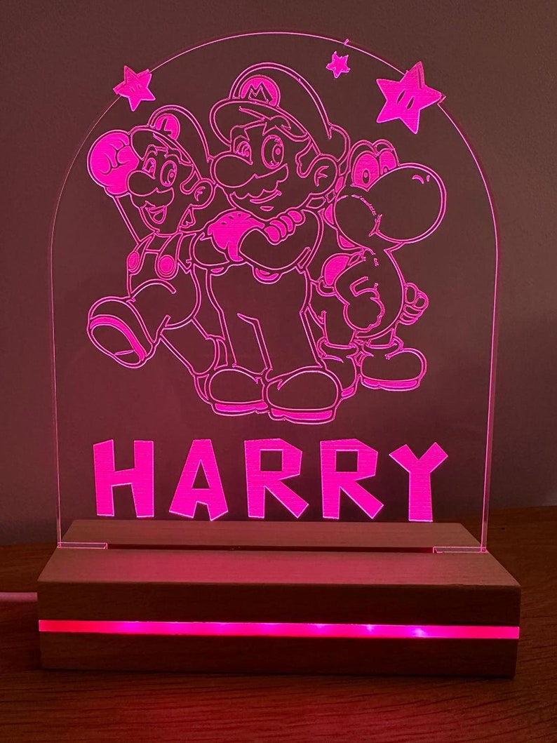 May include: A pink acrylic night light with a wood base. The light features a silhouette of Mario, Luigi, and Yoshi from the Super Mario Bros. video game series. The name "HARRY" is written below the characters.