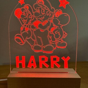May include: A red illuminated acrylic night light featuring Mario, Luigi, and Yoshi characters from the Super Mario Bros. video game series. The name "HARRY" is written below the characters. The night light is on a wooden base.