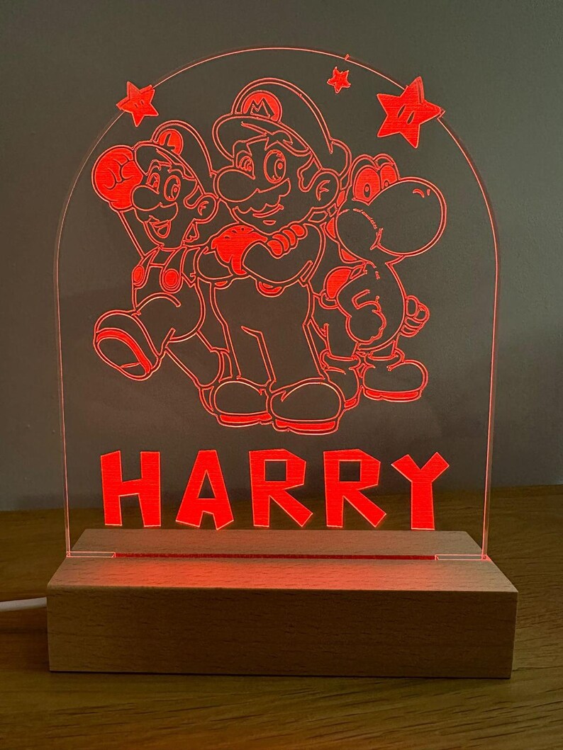 May include: A red illuminated acrylic night light featuring Mario, Luigi, and Yoshi characters from the Super Mario Bros. video game series. The name "HARRY" is written below the characters. The night light is on a wooden base.