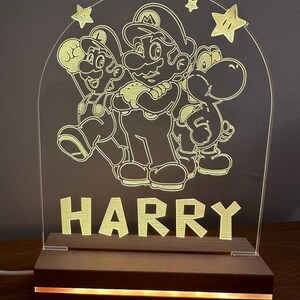 May include: A white acrylic night light with a design of Mario, Luigi, and Yoshi characters from the Super Mario Bros. video game series. The light is illuminated with a warm yellow glow and has the name "HARRY" written below the characters. The light sits on a wooden base with a warm yellow LED light strip.