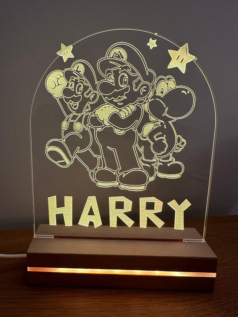 May include: A white acrylic night light with a design of Mario, Luigi, and Yoshi characters from the Super Mario Bros. video game series. The light is illuminated with a warm yellow glow and has the name "HARRY" written below the characters. The light sits on a wooden base with a warm yellow LED light strip.
