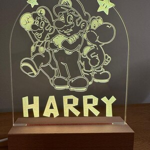 May include: A wooden night light with a clear acrylic panel featuring a silhouette of Mario, Luigi, and Yoshi from the Super Mario Bros. video game series. The name "HARRY" is printed below the characters.