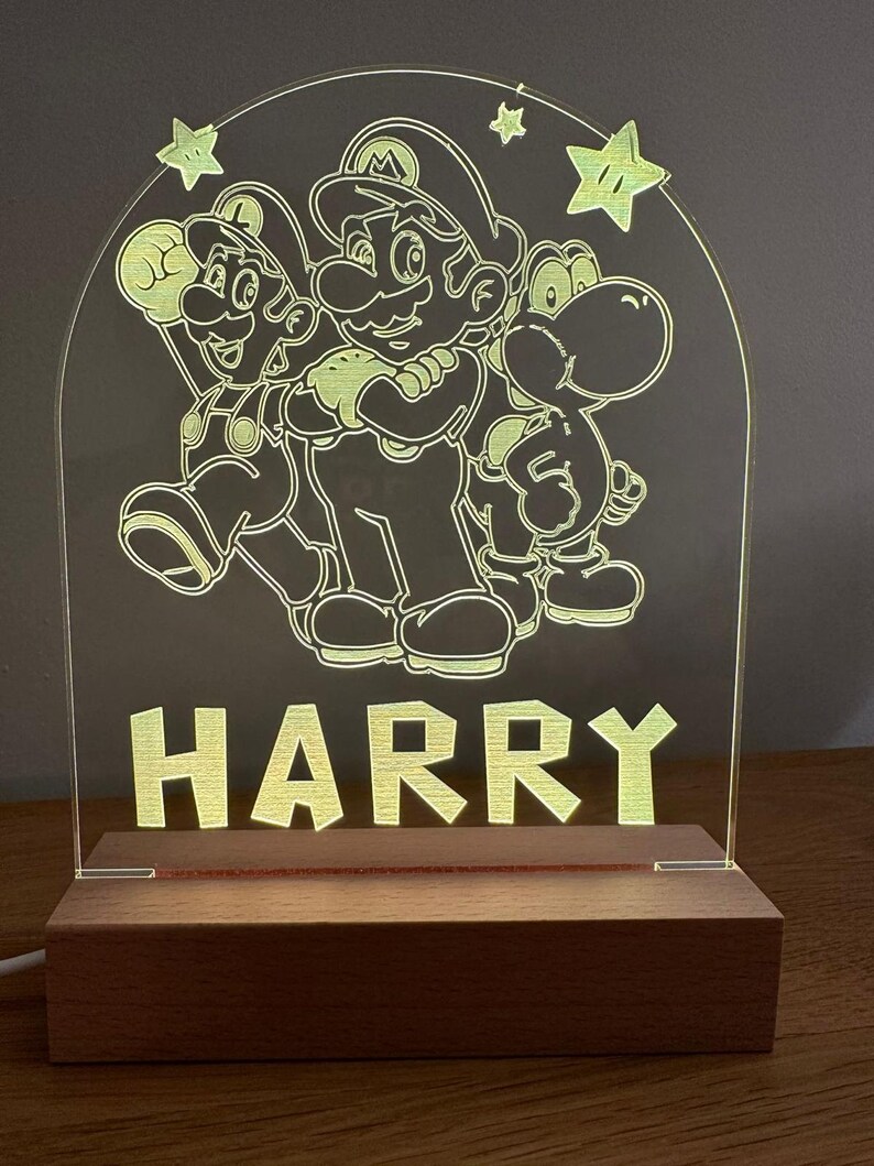 May include: A wooden night light with a clear acrylic panel featuring a silhouette of Mario, Luigi, and Yoshi from the Super Mario Bros. video game series. The name "HARRY" is printed below the characters.
