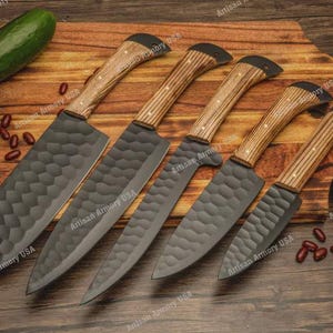 May include: Five kitchen knives with wooden handles and textured dark gray blades are arranged on a wooden cutting board. A green bell pepper, cucumber, and tomatoes are also present. The knives are for food preparation.