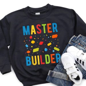 May include: Black sweatshirt with the words "MASTER BUILDER" in colorful block letters. The sweatshirt is paired with blue jeans and white sneakers with black stripes.