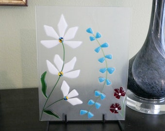 May include: A white frosted glass panel with a floral design. The design features white flowers with yellow centers and blue flowers. The panel is on a black stand.