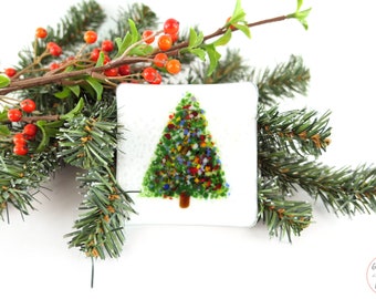 May include: A square, white glass ornament with a fused glass Christmas tree design. The tree is green with colorful accents. The ornament is surrounded by evergreen branches and red berries, creating a festive holiday theme.