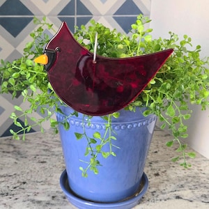 May include: A stained glass cardinal bird decoration in shades of red, black, and yellow, hanging above a blue ceramic pot filled with green foliage. The bird has a metal wire for hanging. The pot sits on a matching saucer.