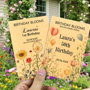 May include: Two rectangular seed packets made of light tan paper. The left packet reads BIRTHDAY BLOOMS, Laura's 1st Birthday, and 05.06.2025. The right packet reads BIRTHDAY BLOOMS WILDFLOWER SEEDS, Laura's 50th Birthday, and 05.06.2025. Both feature floral illustrations.