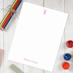 May include: A white notepad with a pink letter L monogram and the name "Rebecca Lopez" printed at the bottom. The notepad is on a white wooden surface with colored pencils and paint pots.
