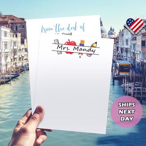 Pu&ograve; includere: Blocco note bianco con un bordo blu e nero e il testo "from the desk of... David" e "Mrs. Mandy". Il blocco note ha una mela rossa, una matita, un righello e una gomma da cancellare.