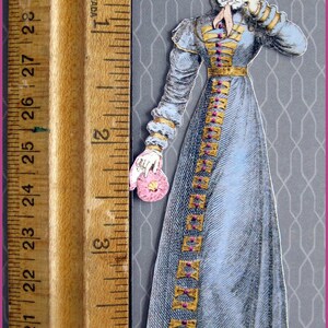 May include: A vintage paper doll of a woman wearing a blue dress with gold trim and a pink and white hat. The doll is holding a pink flower.