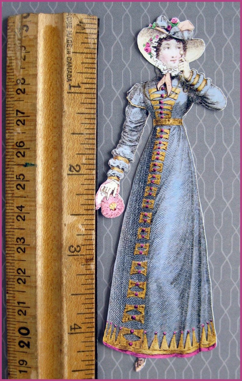 May include: A vintage paper doll of a woman wearing a blue dress with gold trim and a pink and white hat. The doll is holding a pink flower.