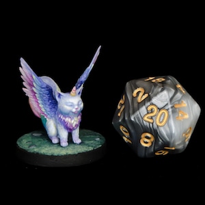 Puede incluir: Una figurita de gato alado con un cuerno de unicornio, pintada en tonos de morado, rosa y azul, se encuentra sobre una base verde. Un dado negro y blanco de 20 caras con números dorados está en el fondo.