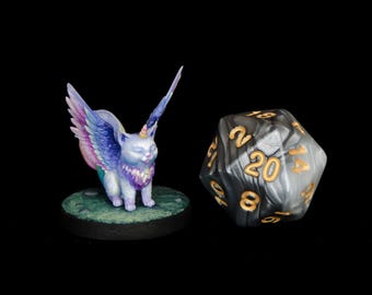 Könnte beinhalten: Eine geflügelte Katzenfigur mit einem Einhornhorn, in Schattierungen von Lila, Rosa und Blau bemalt, steht auf einem grünen Sockel. Ein schwarz-weißer 20-seitiger Würfel mit goldenen Zahlen befindet sich im Hintergrund.