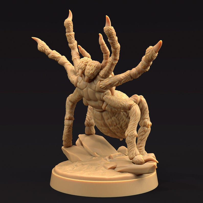 May include: A beige, detailed figurine of a large spider with multiple legs and sharp claws. The spider is posed on a small rock formation, mounted on a circular base. The figurine has a textured, furry appearance.