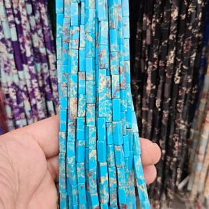 May include: Close-up of strands of turquoise rectangular beads with brown and tan veining. The beads are held in a hand, with other strands of beads in purple and black visible in the background. These beads are suitable for jewelry making.