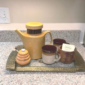 May include: A brown and yellow ceramic teapot with a brown and white ceramic honey pot and four brown and white ceramic mugs on a gold tray. The honey pot has a bee on top of it. The tray has a textured surface.