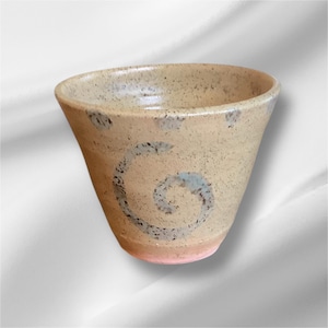 May include: A handmade ceramic cup with a speckled beige exterior and a pink base. The cup features a blue and black spiral design on the side. The cup has a wide opening and a tapered shape.