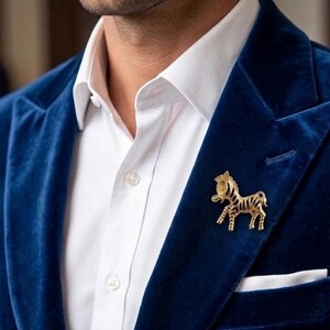 May include: A gold-colored zebra brooch pinned to a dark blue velvet blazer. The brooch is detailed with black stripes and a crown. The blazer is worn over a white dress shirt with a pocket square.