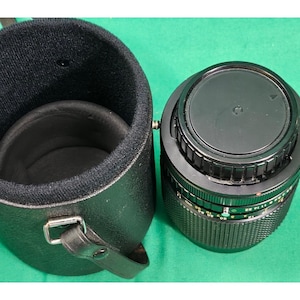 May include: A black camera lens with a textured grip and visible aperture markings alongside a matching black cylindrical carrying case with a soft interior lining and a sturdy metal strap attachment point for secure transport.