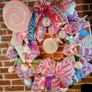 May include: A pink and white candy-themed Christmas wreath with a large pink lollipop, a white pom-pom, and various candy-themed ornaments. The wreath is decorated with pink and white striped mesh and blue and white polka dot ribbon.
