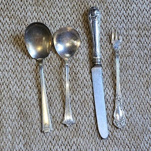 May include: A set of four silver-toned utensils including a large oval spoon, a smaller round spoon, a dinner knife with an ornate handle, and a small three-pronged fork. Each piece features metallic surfaces with visible signs of age.