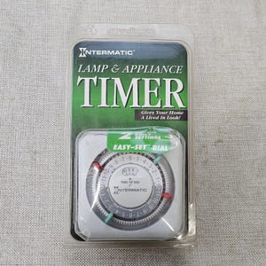 Könnte beinhalten: Eine Intermatic Lampen- und Geräteschaltuhr in transparenter Kunststoffverpackung. Die weiße, quadratische Zeitschaltuhr verfügt über ein rundes Zifferblatt mit AM- und PM-Markierungen, eine zentrale Zeitanzeige sowie zwei rote und zwei türkise Stifte.