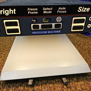 Peut inclure: Un agrandisseur vidéo électronique Freedom Machine doté d'un panneau de commande noir avec des étiquettes en texte blanc, notamment Bright, Freeze Frame, Select Mode, Auto Focus et Size. L'appareil possède des boutons noirs, des icônes et une base blanche.