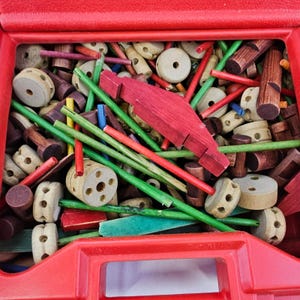May include: A red plastic carrying case filled with a collection of wooden construction toy pieces, including natural wood wheels with holes, various lengths of green, red, and purple painted wooden dowels, and a large red wooden block.