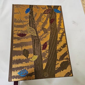 May include: A hardcover book featuring a gold and brown background with two dark brown tree trunks and scattered leaves in shades of blue, yellow, and maroon. The cover design includes a textured pattern and a dark brown spine with a ribbon bookmark.