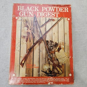 May include: A vintage paperback book titled Black Powder Gun Digest, edited by Toby Bridges. The red cover features an illustration of two crossed muzzleloading rifles, a small pistol, a raccoon pelt, a leather pouch, and a ceramic jug.