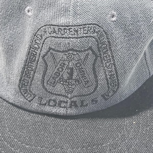 May include: A dark grey baseball cap featuring a black embroidered emblem on the front. The emblem includes the text UNITED BROTHERHOOD OF CARPENTERS AND JOINERS OF AMERICA, LABOR OMNIA VINCIT, and LOCAL 5, with a central shield and tool design.