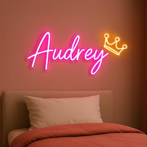 Puede incluir: Un letrero de ne&oacute;n rosa con el nombre "Audrey" y una corona amarilla de ne&oacute;n. El letrero est&aacute; montado en una pared rosa sobre una cama con una almohada blanca y una manta roja.