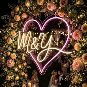 May include: A neon sign in the shape of a heart with the letters "M&Y" in a cursive font. The sign is surrounded by a floral arrangement of roses and greenery, with string lights. The background is dark, suggesting a nighttime event.
