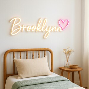 Puede incluir: Una cabecera de cama de madera blanca con un letrero de ne&oacute;n blanco que dice "Brooklynn" con un coraz&oacute;n de ne&oacute;n rosa. La cama tiene una almohada blanca y una colcha verde.