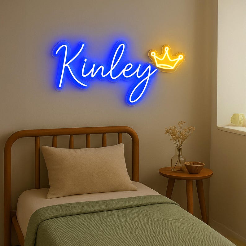 Puede incluir: Un letrero de ne&oacute;n azul con el nombre "Kinley" y una corona de ne&oacute;n amarilla. El letrero est&aacute; montado en una pared blanca sobre una cama con una manta verde.