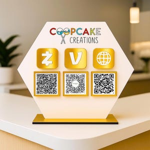 May include: A gold-colored display stand with the text "COOPCAKE CREATIONS" and three icons above three QR codes. The icons include a stylized "Z", a "V", and a globe. The stand is on a white surface.