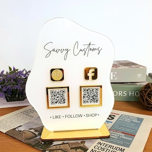 May include: A white acrylic display stand with gold detailing, inscribed with "Savvy Customs" in black script. It features gold-framed icons for a globe and Facebook, QR codes, and the text "LIKE • FOLLOW • SHOP."