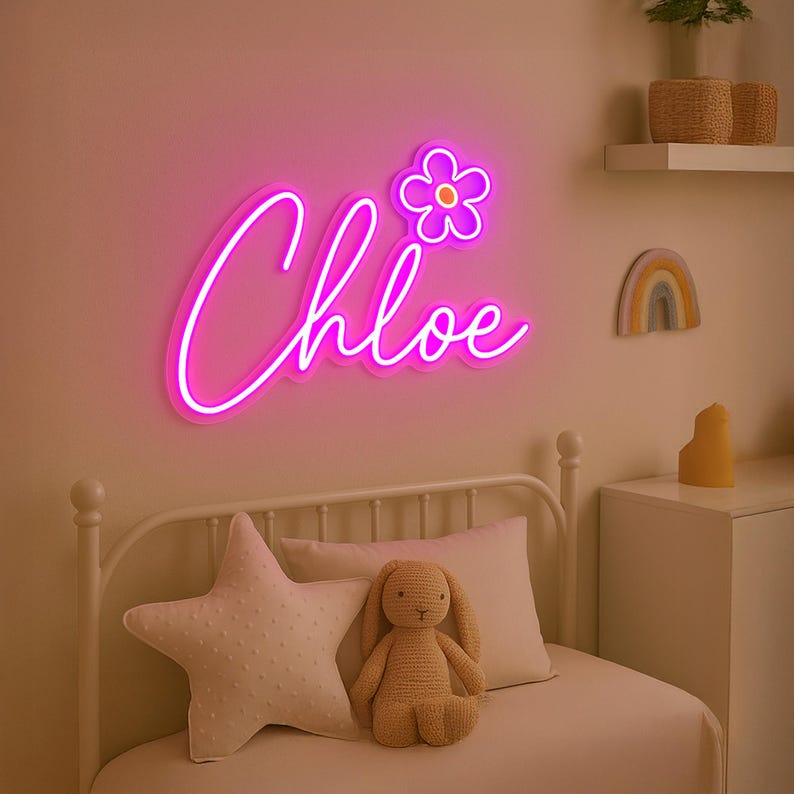 Puede incluir: Un letrero de ne&oacute;n rosa con el nombre "Chloe" en una fuente cursiva con una flor rosa encima. El letrero est&aacute; montado en una pared blanca sobre una cama blanca con una almohada blanca en forma de estrella y un conejito de peluche marr&oacute;n.