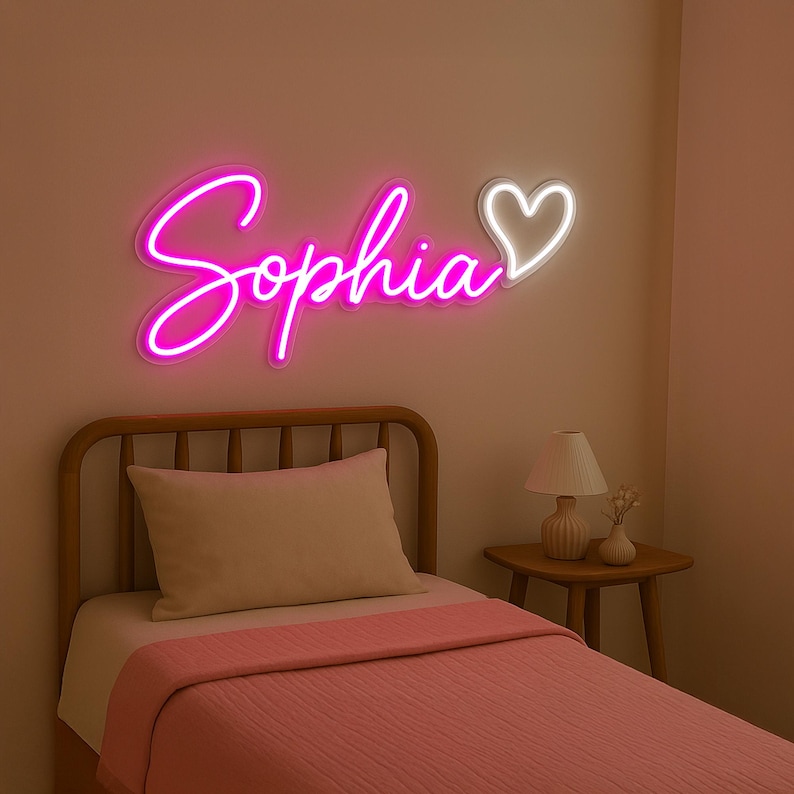 Puede incluir: Un letrero de ne&oacute;n rosa con el nombre "Sophia" y un coraz&oacute;n blanco. El letrero est&aacute; montado en una pared blanca sobre un marco de cama de madera con una manta rosa.