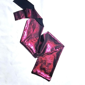 Puede incluir: Un cinturón fucsia metalizado con la parte inferior negra, sobre una superficie blanca. El cinturón está doblado en zigzag, mostrando su superficie brillante y texturizada. Los extremos del cinturón son rectangulares.