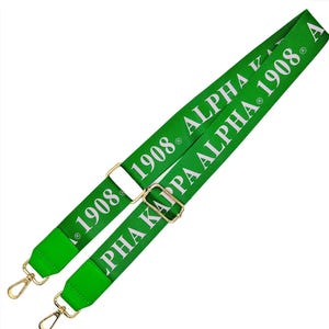 May include: A green adjustable bag strap with gold-colored metal hardware. The strap features the text "ALPHA KAPPA ALPHA 1908" in white. The strap has a solid green color with white text.
