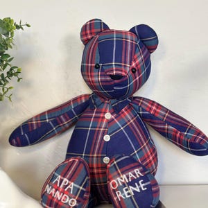 May include: A handmade teddy bear with a blue, red, and yellow plaid pattern. The bear has button details and embroidered text on its feet. The bear is sitting with its arms outstretched.