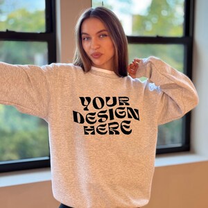 May include: A gray sweatshirt with the text "YOUR DESIGN HERE" in black. The sweatshirt is being worn by a person with long brown hair.