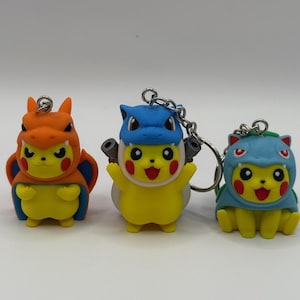 May include: Three colorful Pikachu keychains, each dressed in a different costume. The keychains are yellow with red cheeks and black eyes. One is in an orange dragon costume, another in a blue and white costume, and the third in a blue costume.