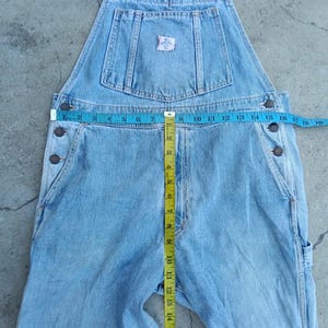 Op de afbeelding: Lichtblauwe denim overall met verstelbare bandjes, meerdere zakken en knoopsluitingen. Een meetlint geeft aan dat de taille ongeveer 35,5 cm breed is. De overall heeft een kleine patch op de borstzak.